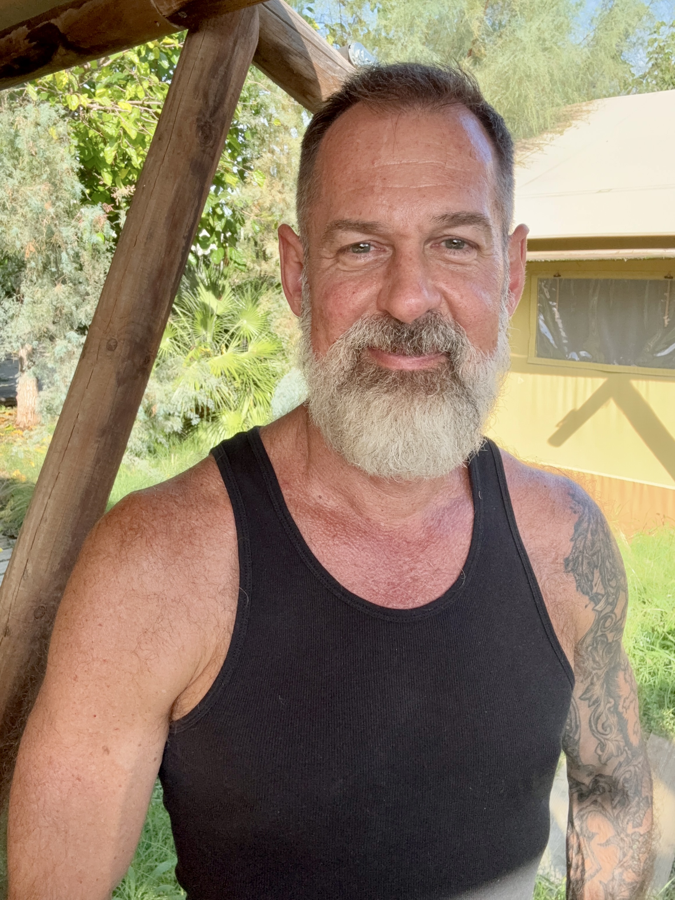 Alex Sloan outdoors in a black tank top, smiling, with timber beams and a yellow cabin behind him